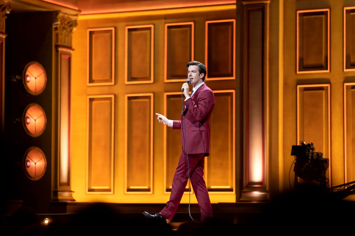 Un'inquadratura ampia del comico John Mulaney mentre cammina sul palco proprio su uno sfondo arancione con un abito rosso nel suo speciale Netflix Baby J.
