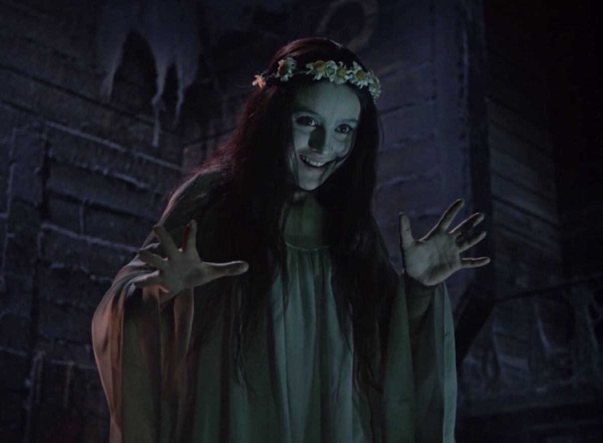 Una donna sorridente dal viso pallido che indossa una corona di fiori e un vestito verde gesticola con le mani aperte in Viy.