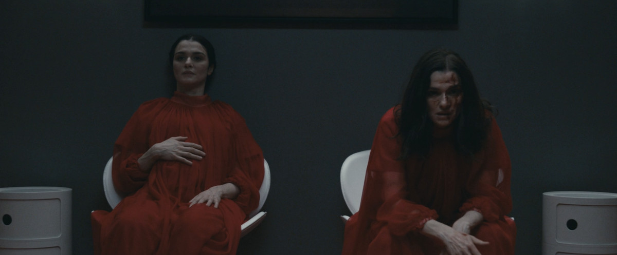 Beverly (Rachel Weisz) ed Elliot (Rachel Weisz) siedono nei loro abiti chirurgici rossi, con Beverly che si stringe la pancia incinta ed Elliot seduto e accigliato con una faccia insanguinata