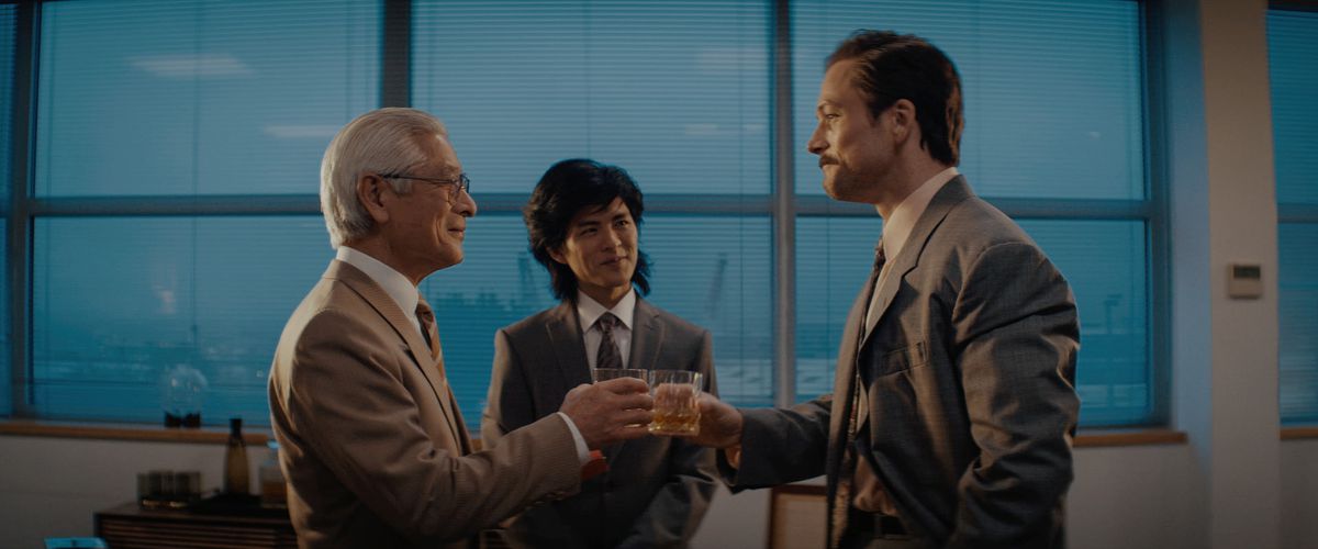 Un anziano giapponese e un giovane occidentale baffuto fanno tintinnare i bicchieri in un ufficio mentre un giovane giapponese con una triglia guarda