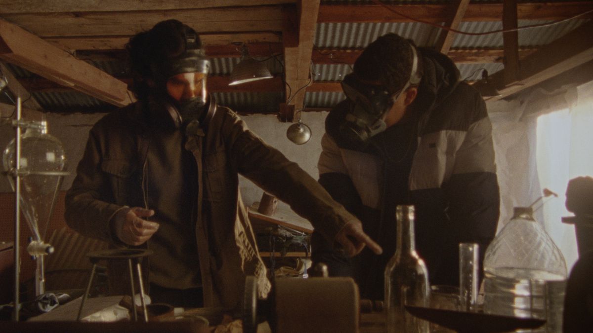 Due persone che indossano maschere antigas lavorano con sostanze chimiche, mentre una indica How to Blow Up a Pipeline.