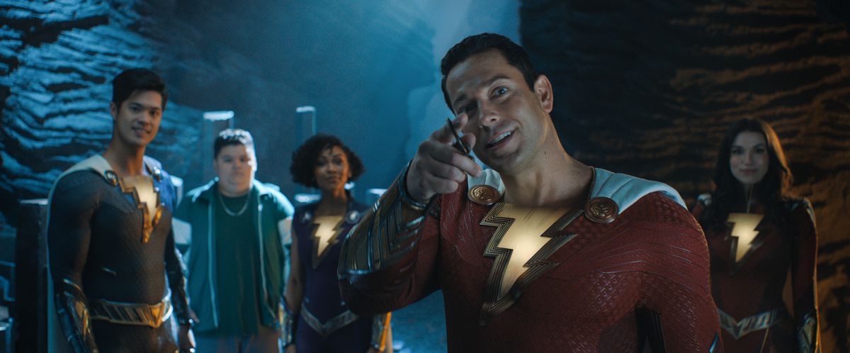 Zachary Levi nei panni di Shazam indica la telecamera e sorride, con i membri della sua famiglia di eroi dietro di lui, in Shazam!  Furia degli Dei