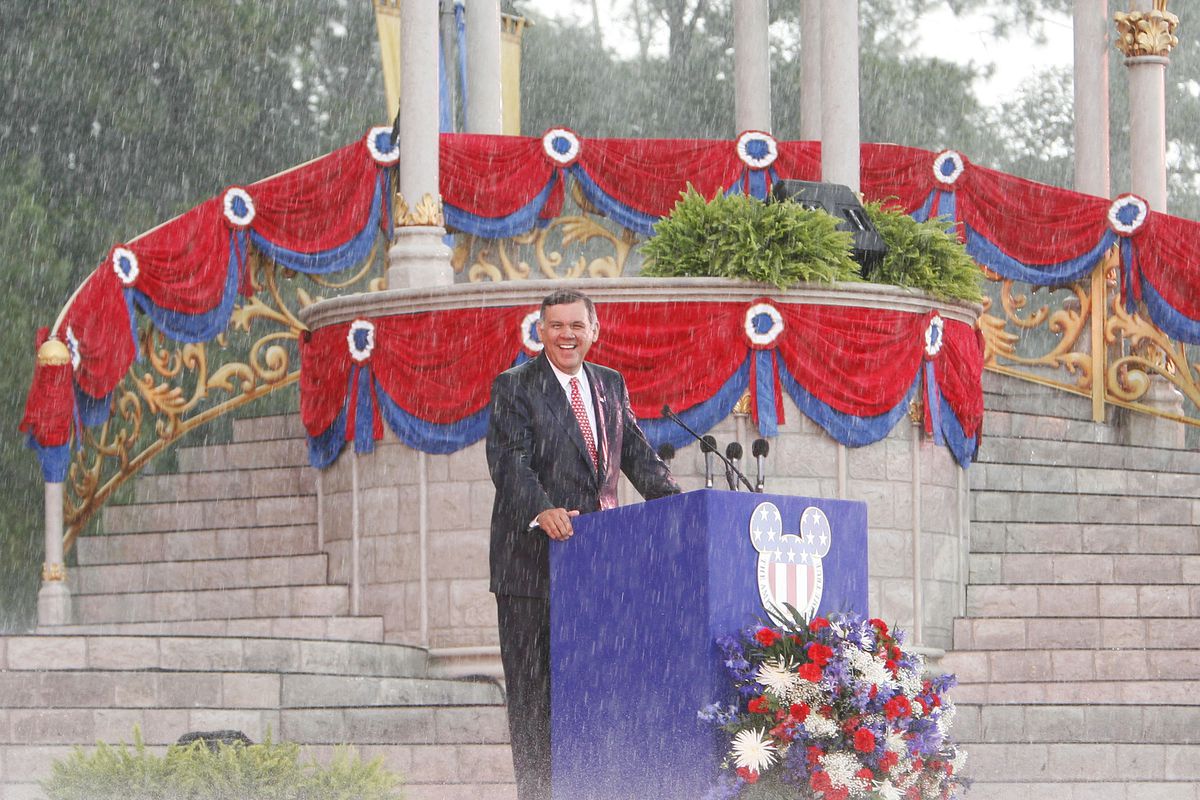 Il senatore degli Stati Uniti Mel Martinez sorride sotto la pioggia battente su un podio all'aperto con una testa di Topolino con sopra la bandiera americana durante una cerimonia di naturalizzazione di massa al Walt Disney World in Florida il 4 luglio 2007