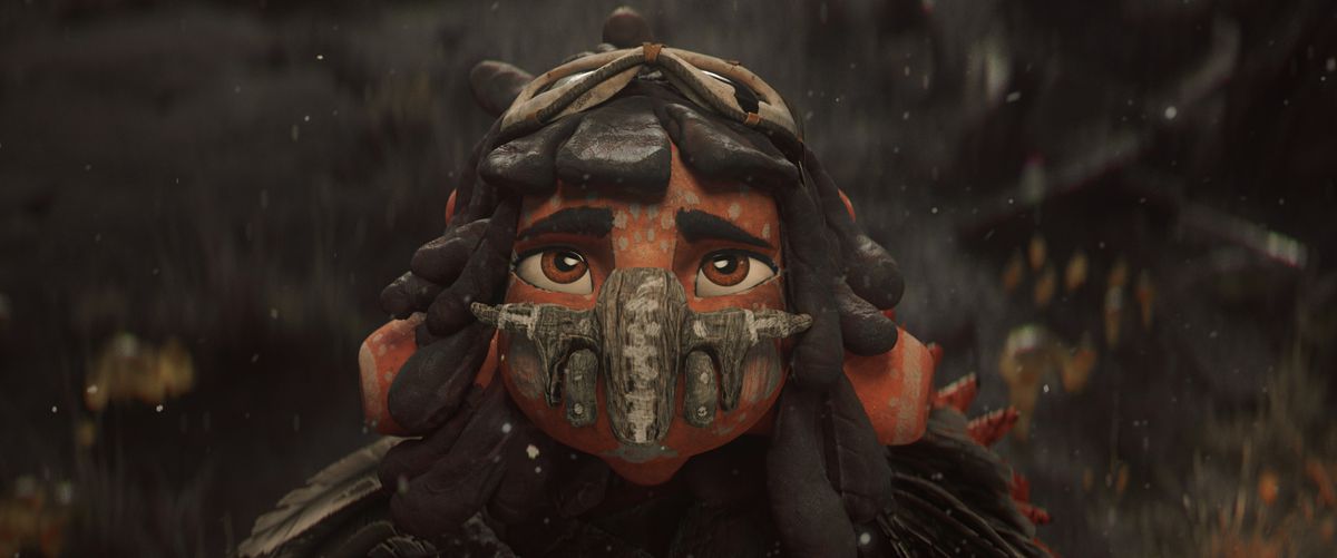 Una giovane ragazza aliena dalla pelle arancione, che indossa una maschera protettiva fatta di pietra e legno.  Alza lo sguardo verso la telecamera, gli occhi castani spalancati per la preoccupazione.