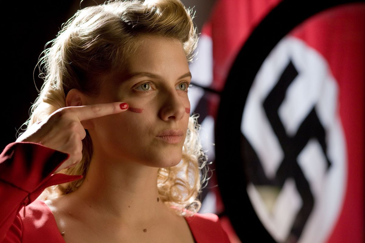 Una giovane donna bionda con un vestito rosso si spalma la pittura di guerra sul viso con una svastica nazista appesa sullo sfondo