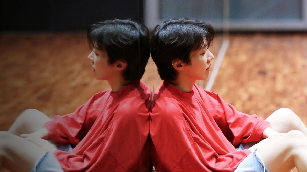 Il rapper dei BTS J-Hope si appoggia a uno specchio lungo il pavimento mentre indossa pantaloncini di jeans e una maglietta a maniche lunghe rosso chiaro.  Vediamo il suo riflesso nello specchio.