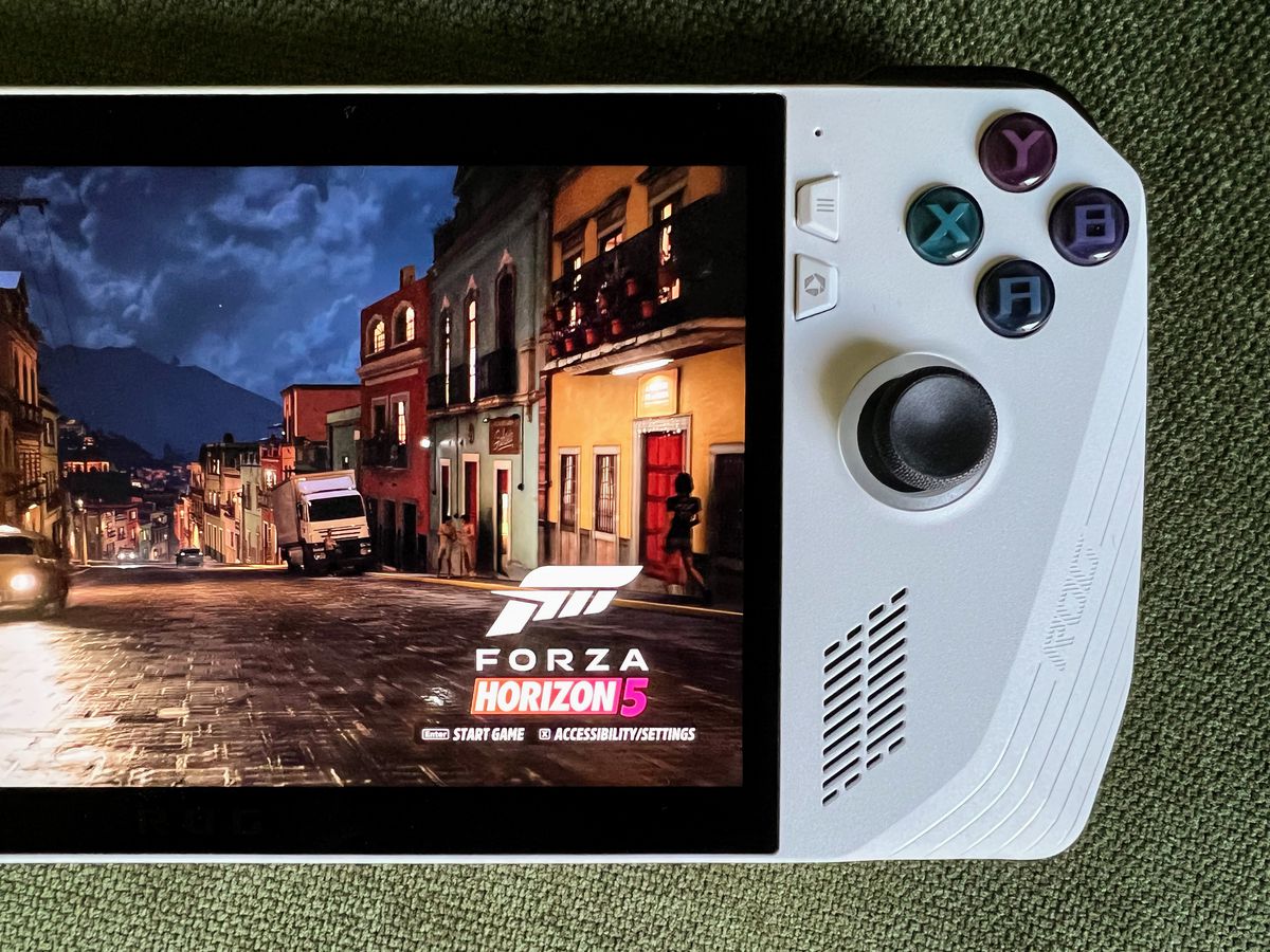 una scena di strada piovosa in Forza Horizon 5 in esecuzione su un palmare da gioco Asus ROG Ally, sdraiato su tessuto verde oliva, in una foto in primo piano dall'alto del lato destro del dispositivo