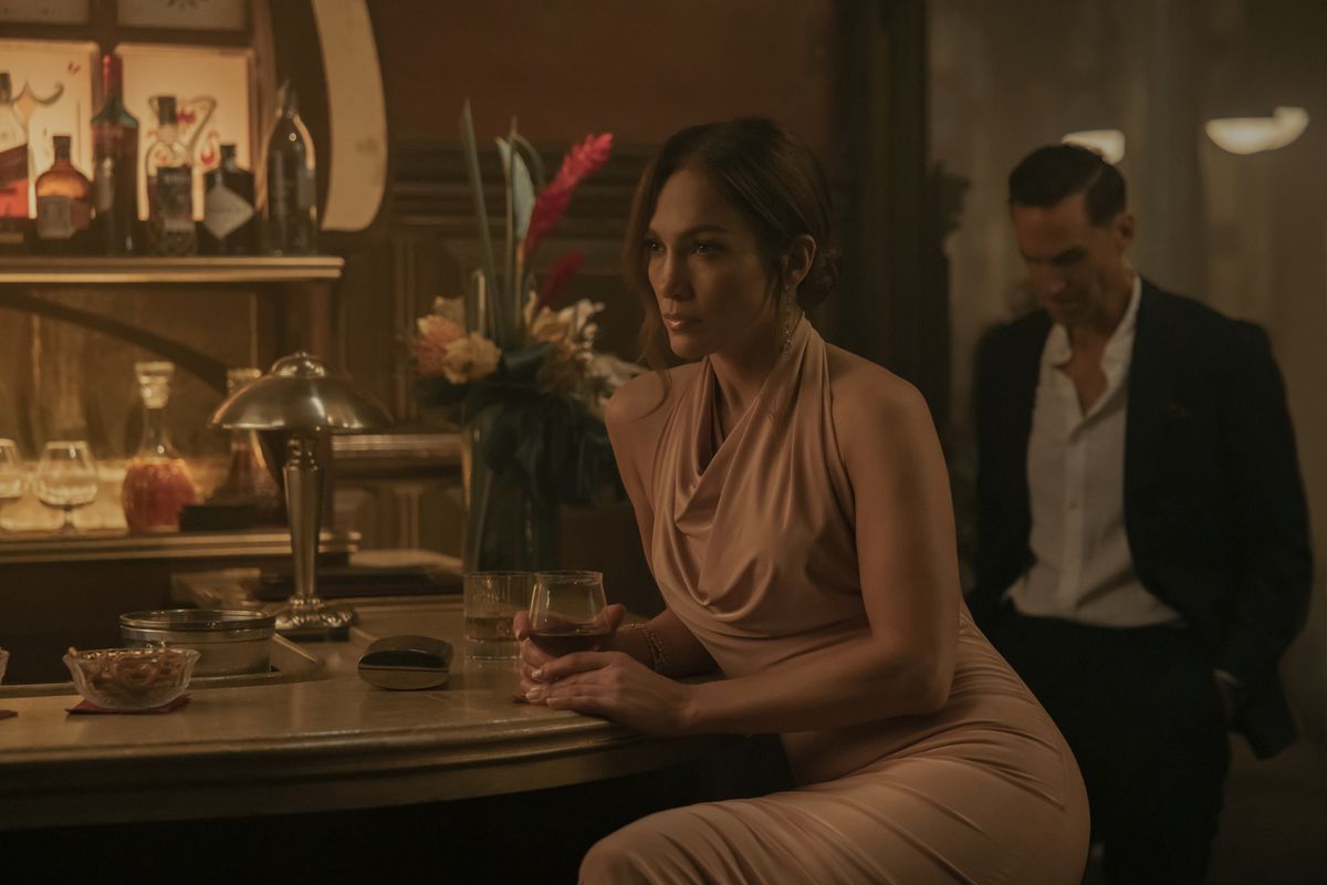 Jennifer Lopez siede in un bar con un bicchiere di vino mentre indossa un bel vestito in The Mother.  Joseph Fiennes incombe dietro di lei, con indosso un completo.