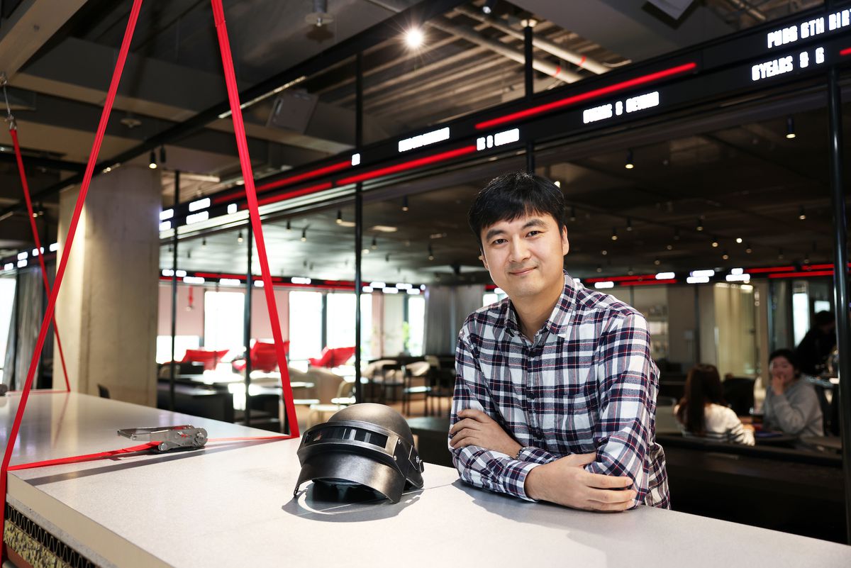 Una foto di Sangkyun Kim di PUBG Studios in piedi, con le braccia incrociate, vicino a un casco PUBG