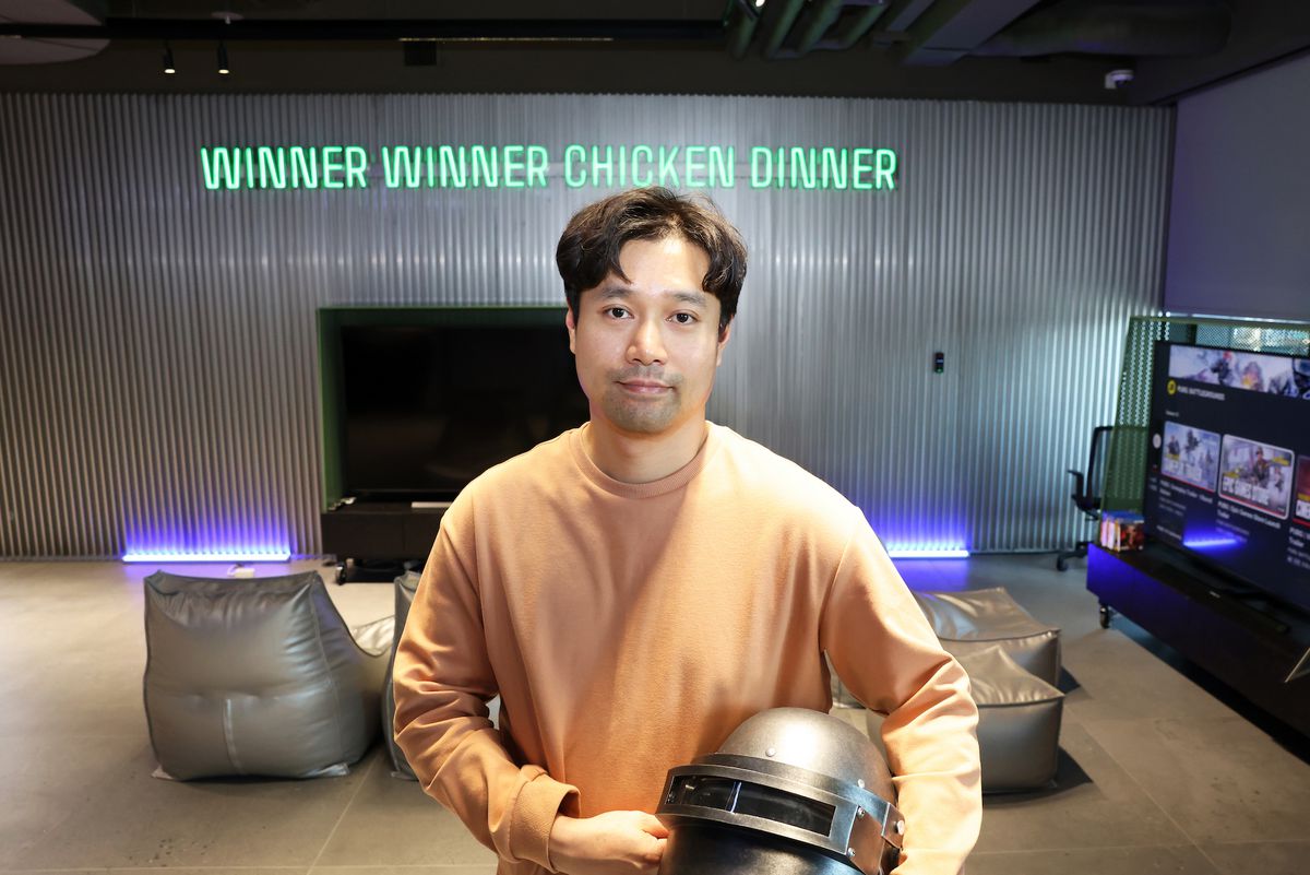 Una fotografia di Taeyhun Kim di PUBG Studios con in mano il classico casco di PUBG e in piedi davanti a un'insegna al neon con la scritta 
