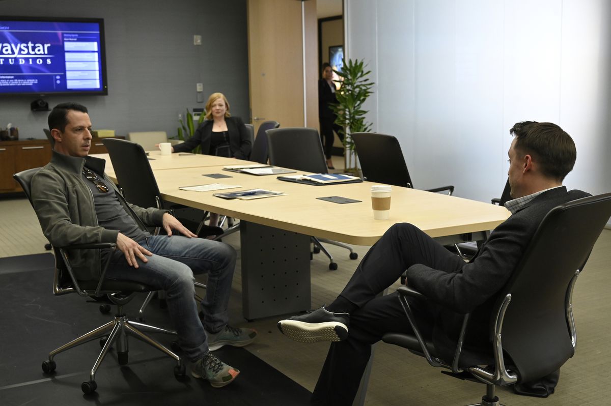 Shiv (Sarah Snook) siede dall'altra parte sullo sfondo a un tavolo da conferenza mentre i suoi fratelli Kendall (Jeremy Strong) e Roman (Kieran Culkin) si siedono e parlano più vicino al primo piano