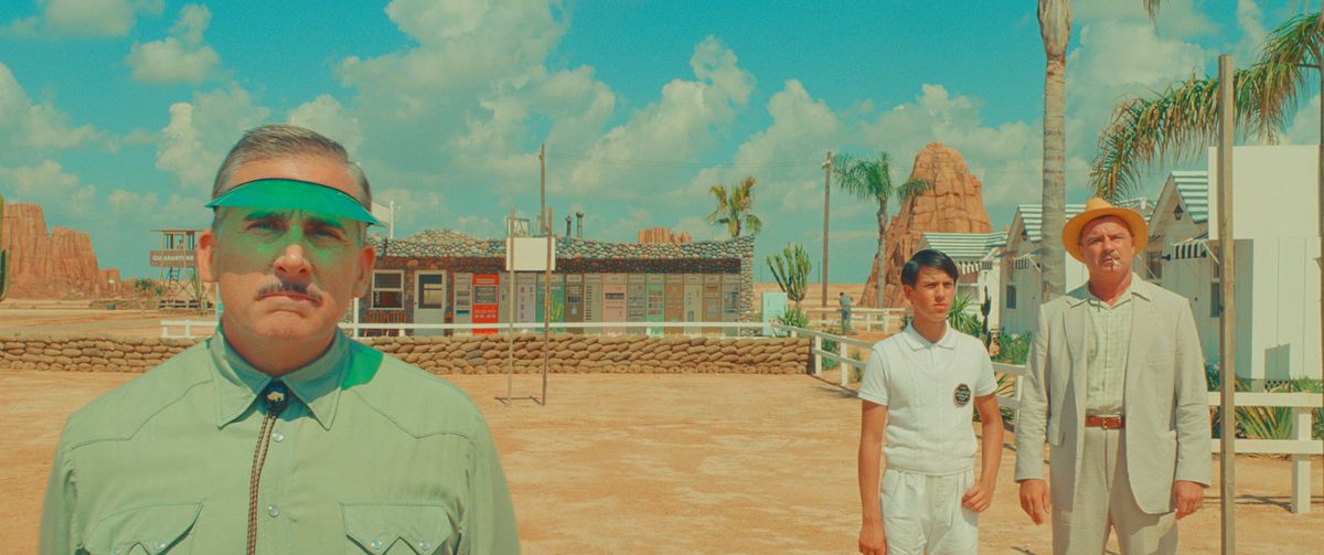 Un manager di un motel (Steve Carell) si trova di fronte a un motel nel deserto, indossa un parasole verde e guarda nella telecamera, mentre due persone in abiti chiari (Aristou Meehan e Liev Schreiber) stanno in lontananza dietro di lui in Asteroid City di Wes Anderson.
