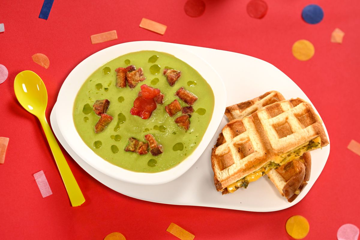Una ciotola di zuppa verde con pezzi galleggianti.  Accanto c'è un panino con i waffle. 