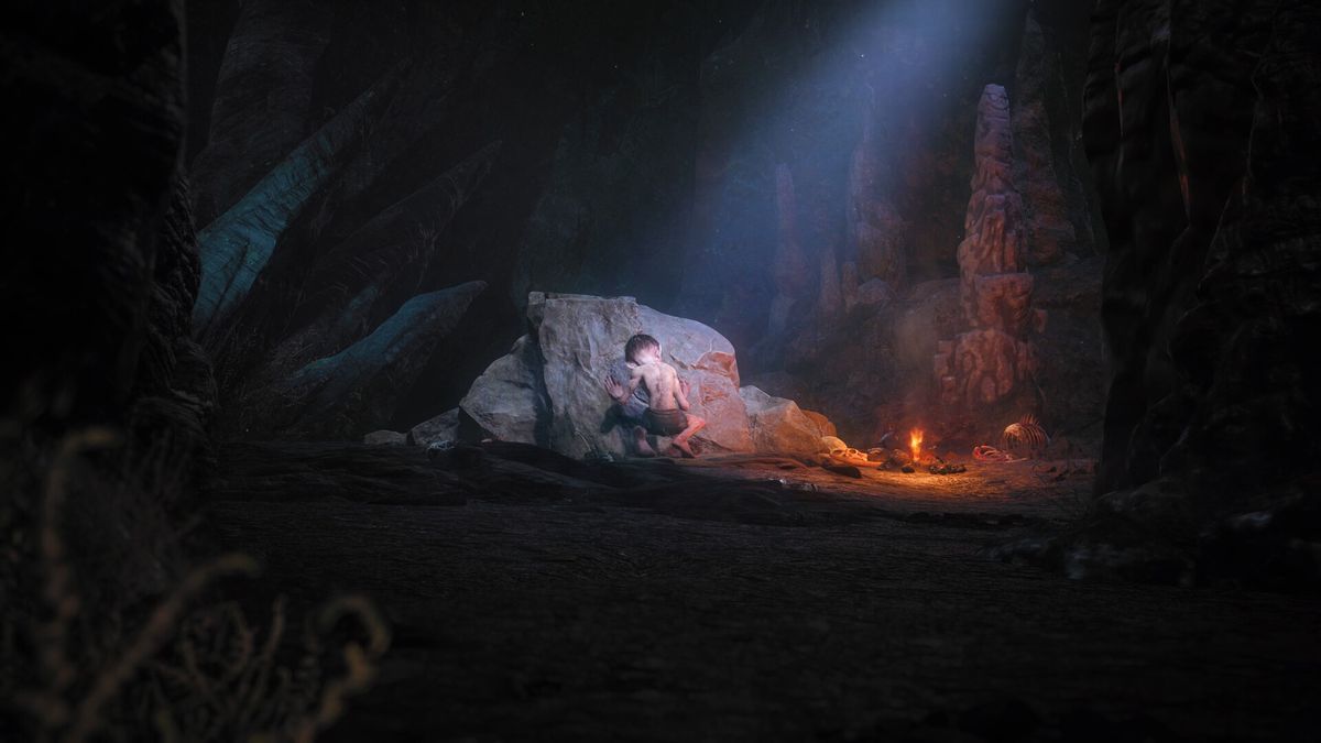 In una caverna buia illuminata da un piccolo fuoco e da un raggio di luce dal cielo, Gollum si accovaccia con le spalle alla 