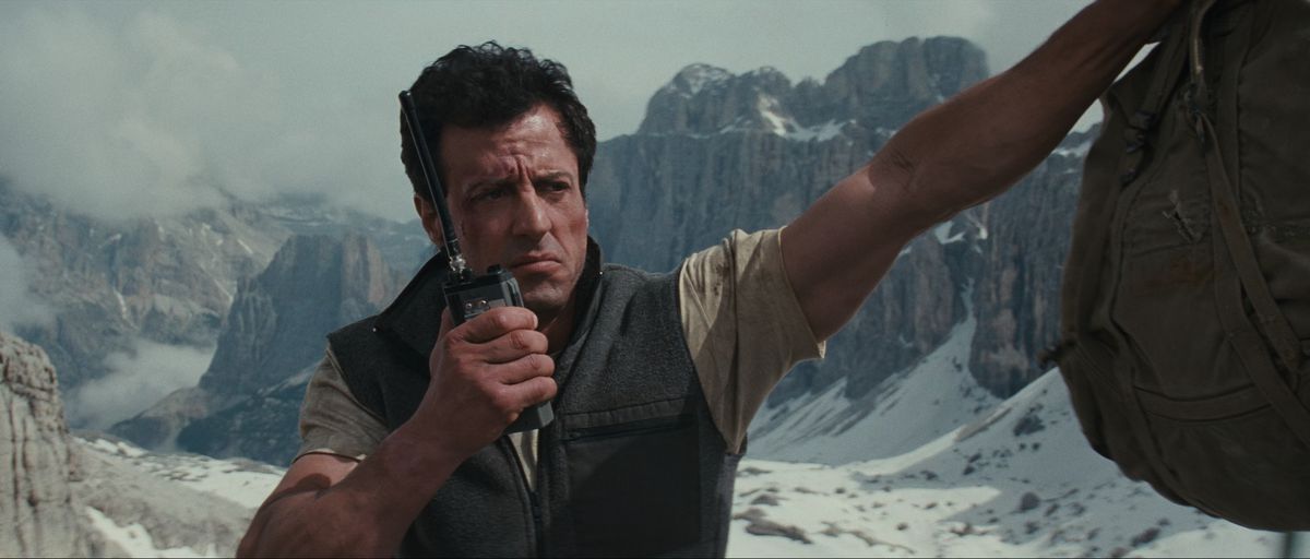 Sylvester Stallone parla alla radio mentre appoggia il braccio su una scogliera in Cliffhanger.  Dietro di lui c'è una splendida vista delle montagne innevate.