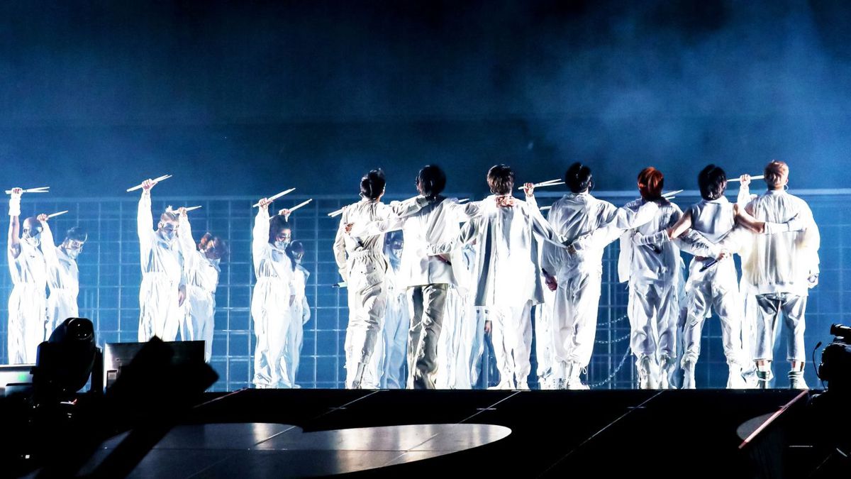I membri dei BTS sono vestiti di bianco, con le braccia l'uno attorno all'altro, in una vista dal retro al loro concerto Permission to Dance on Stage LA.  I ballerini di sottofondo stanno con le braccia in aria.  Tutti tengono le bacchette.