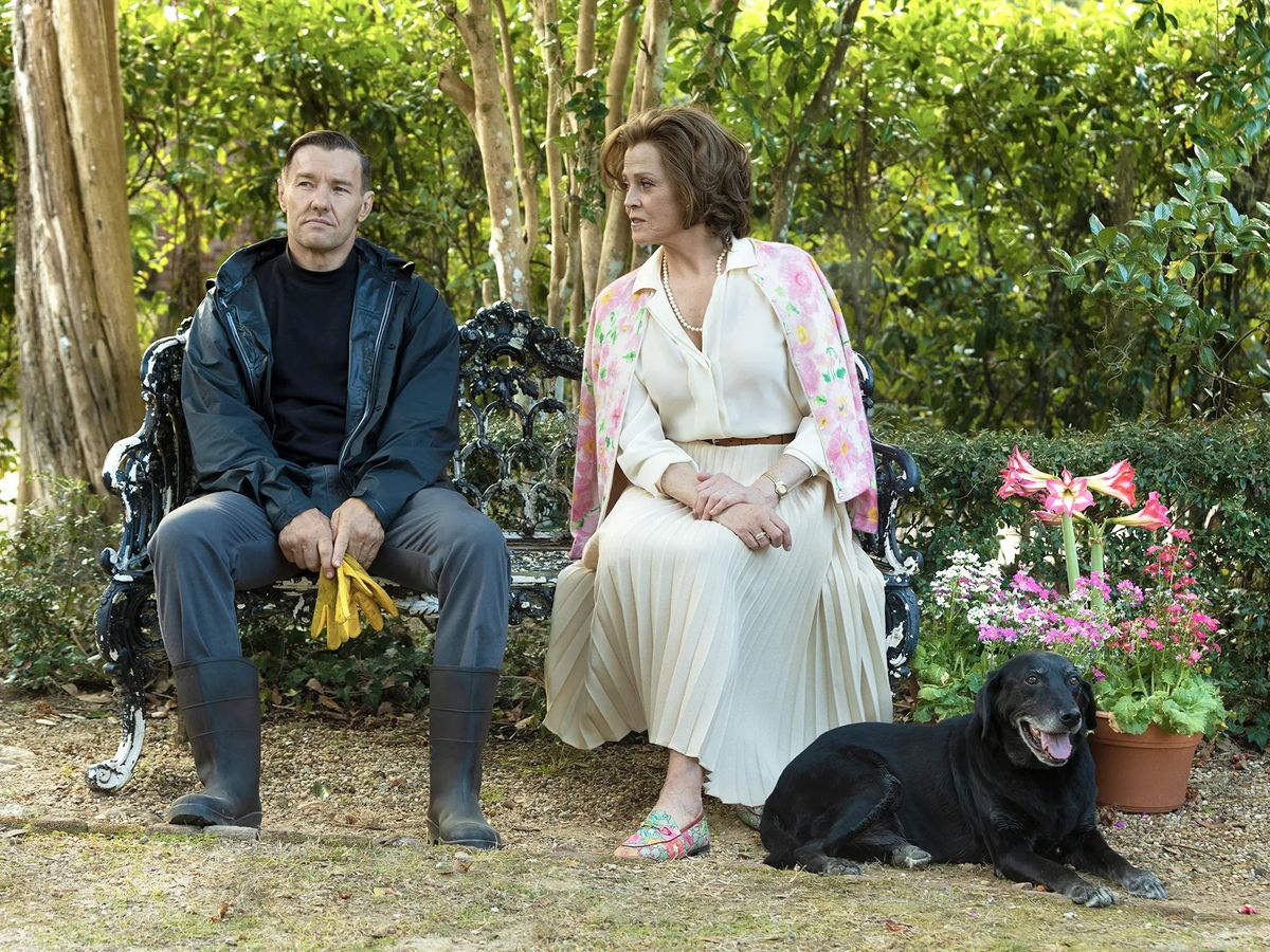 (LR) Joel Edgerton e Sigourney Weaver seduti su una panchina vicino a una composizione di vasi di fiori e un cane nero in Master Giardiniere.