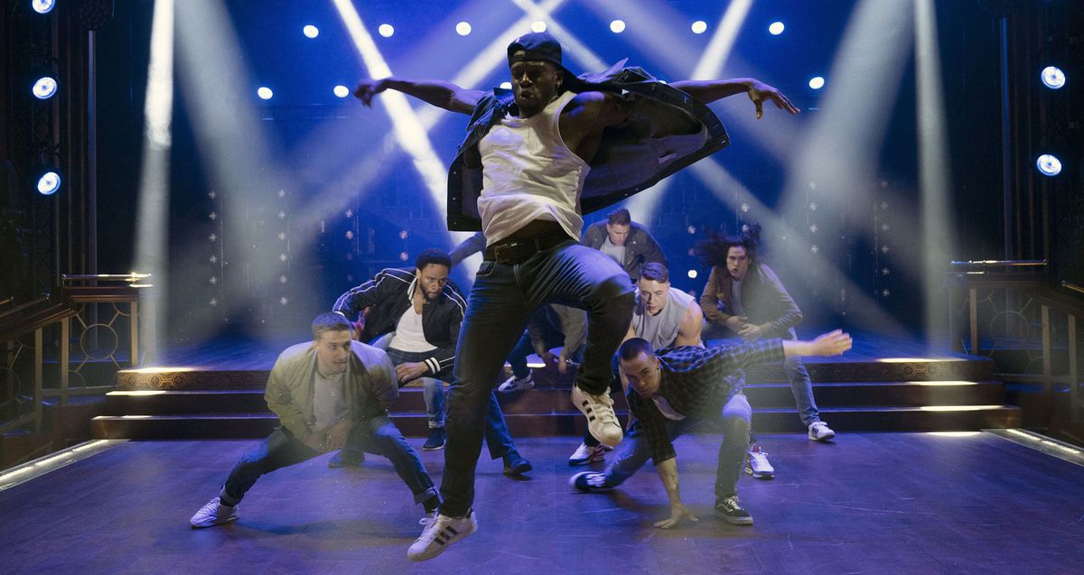 Un ballerino nero in jeans e un berretto da baseball all'indietro salta in aria mentre altri ballerini maschi si accovacciano dietro di lui su un palco illuminato di blu attraversato da luminosi riflettori bianchi in una scena di Magic Mike's Last Dance