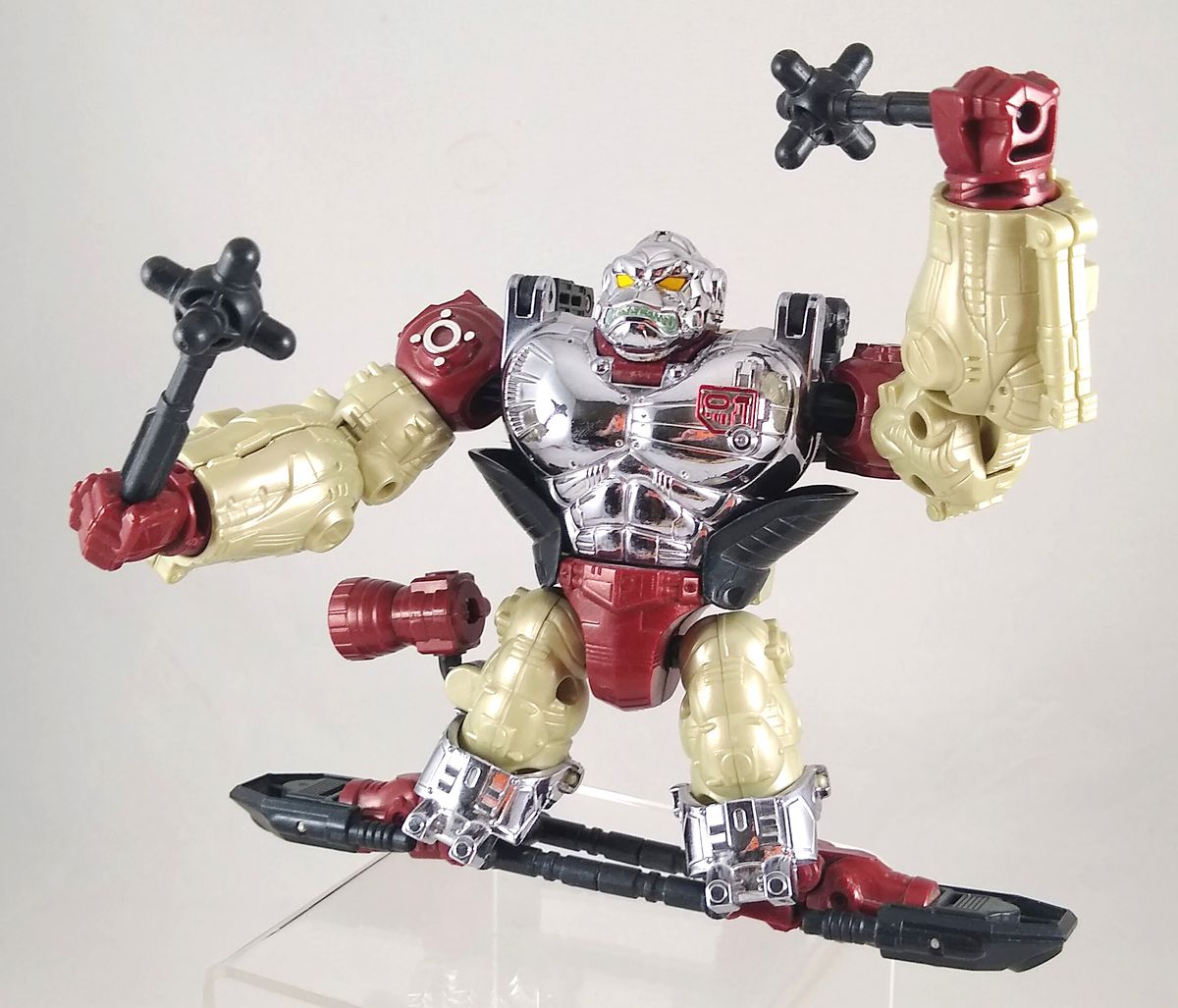 Una foto di un giocattolo Transformers esclusivo della convention Apelinq, una scimmia di metallo rosso, nero, argento e crema in piedi su un hoverboard nero e che brandisce mazze nere